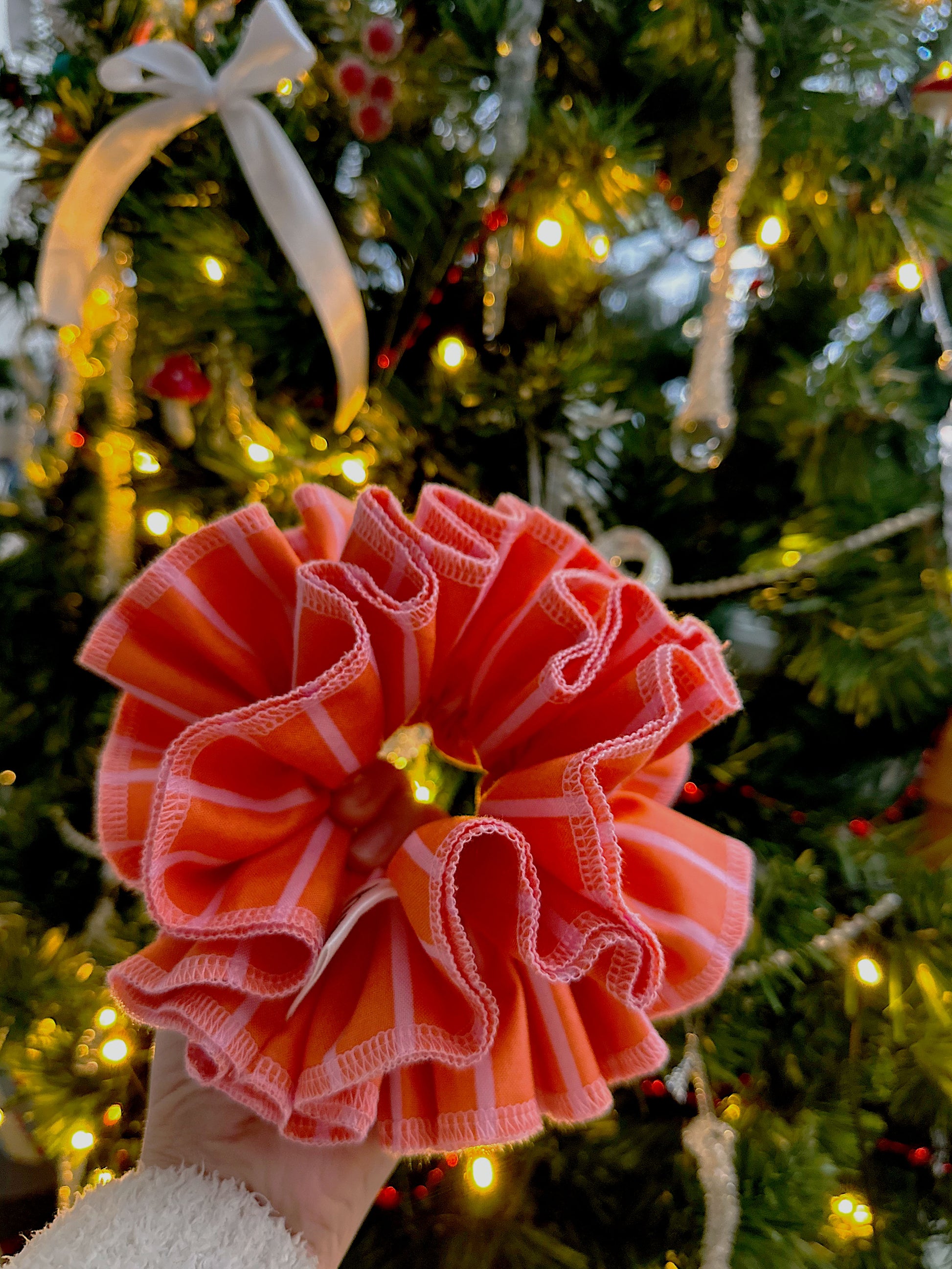 A handmade orange and pink striped cotton scrunchie with contrast pink stitching, displayed in front of c christmas tree.