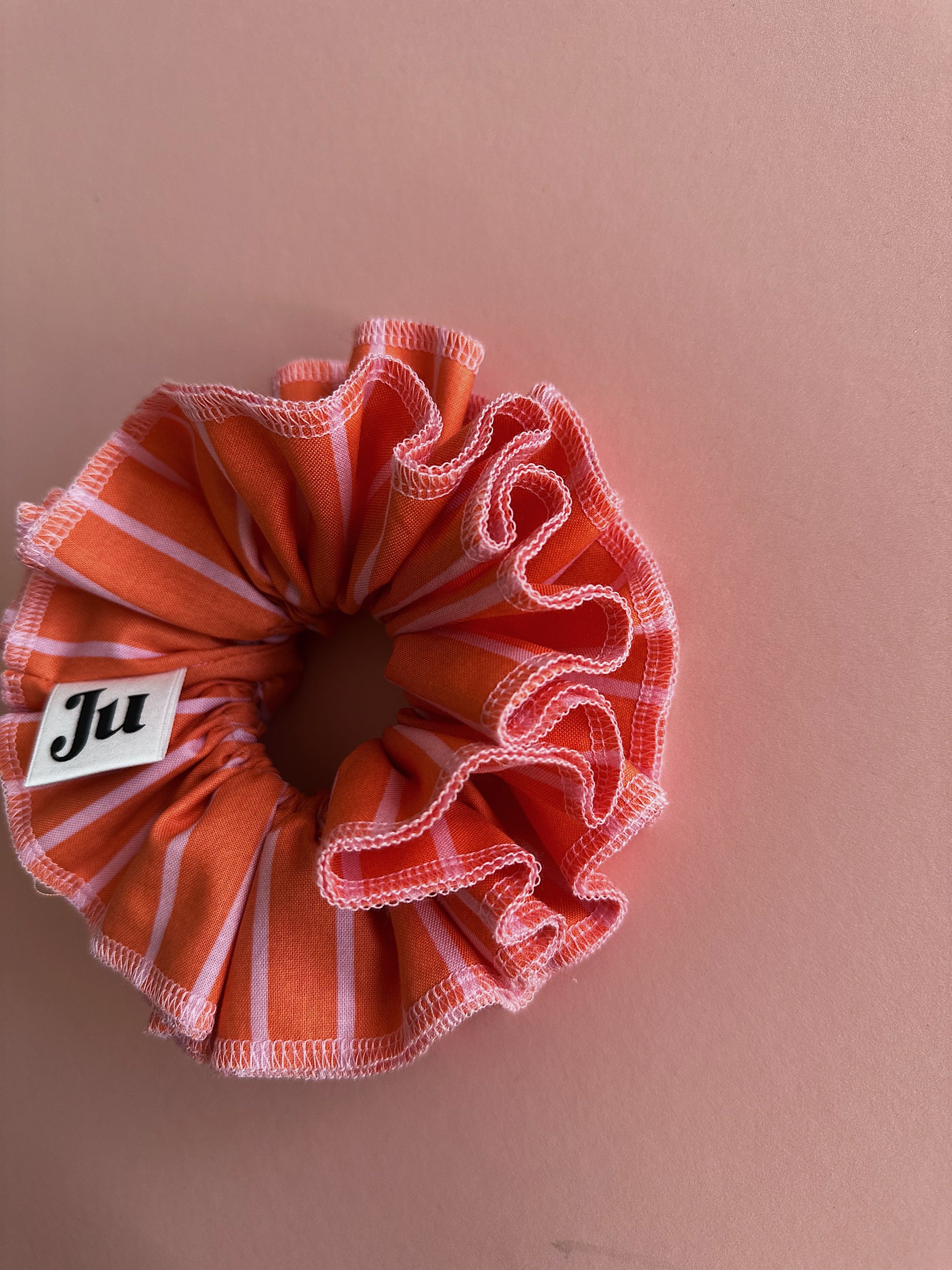 A handmade orange and pink striped cotton scrunchie with contrast pink stitching, displayed on a soft pink background.