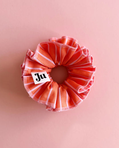 A handmade orange and pink striped cotton scrunchie with contrast pink stitching, displayed on a soft pink background.