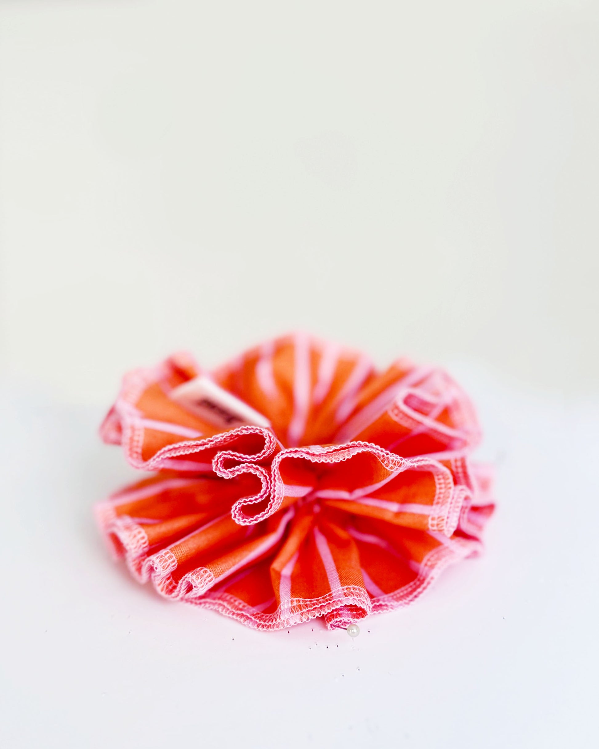 A handmade orange and pink striped cotton scrunchie with contrast pink stitching, displayed on a white  background.