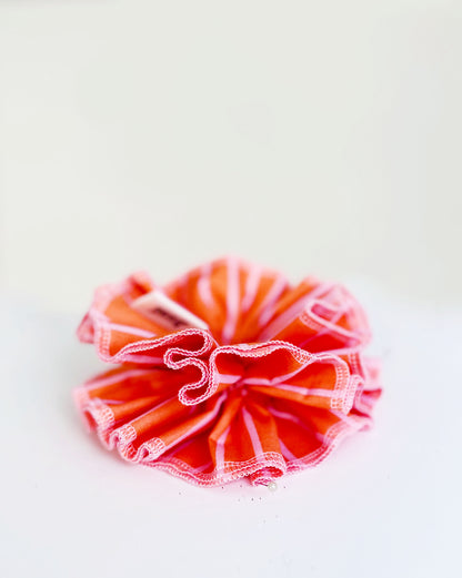 A handmade orange and pink striped cotton scrunchie with contrast pink stitching, displayed on a white  background.