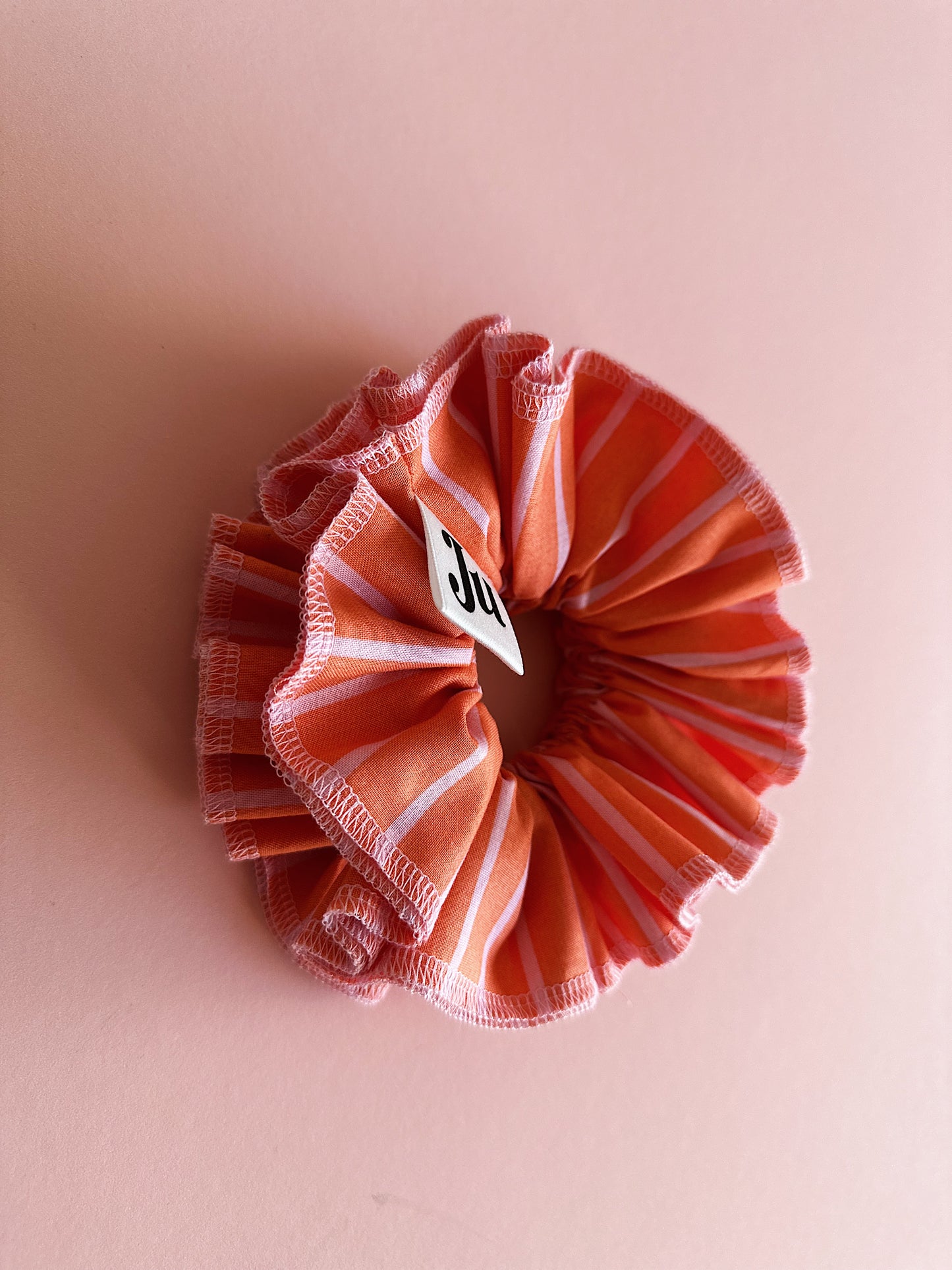 A handmade orange and pink striped cotton scrunchie with contrast pink stitching, displayed on a soft pink background.
