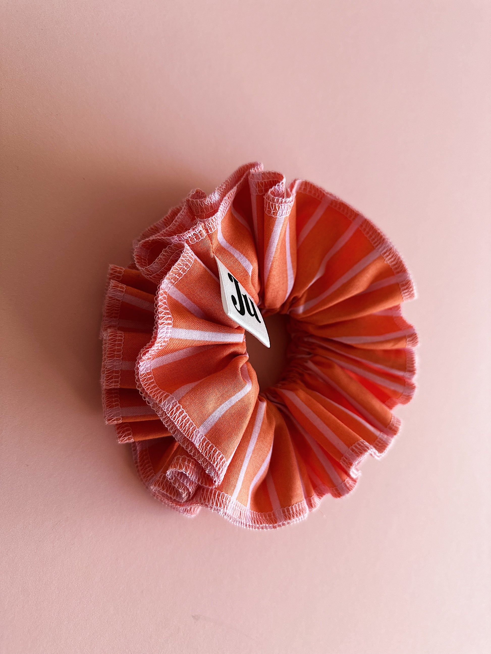 A handmade orange and pink striped cotton scrunchie with contrast pink stitching, displayed on a soft pink background.